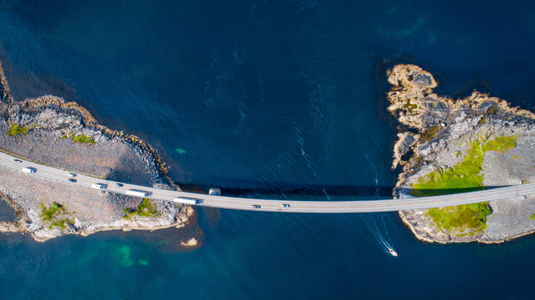 A breathtaking 4K wallpaper showcasing the Storseisundet Bridge, a curving part of Norway's Atlantic Ocean Road, extending gracefully between rocky islands across deep blue water. The deep sapphire tones of the ocean, with a small boat cutting a white wake, beautifully contrast against the stark gray bridge and the rugged, sun-dappled rocky coastline.