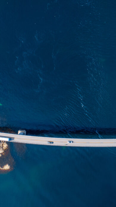 A breathtaking 4K wallpaper showcasing the Storseisundet Bridge, a curving part of Norway's Atlantic Ocean Road, extending gracefully between rocky islands across deep blue water. The deep sapphire tones of the ocean, with a small boat cutting a white wake, beautifully contrast against the stark gray bridge and the rugged, sun-dappled rocky coastline.