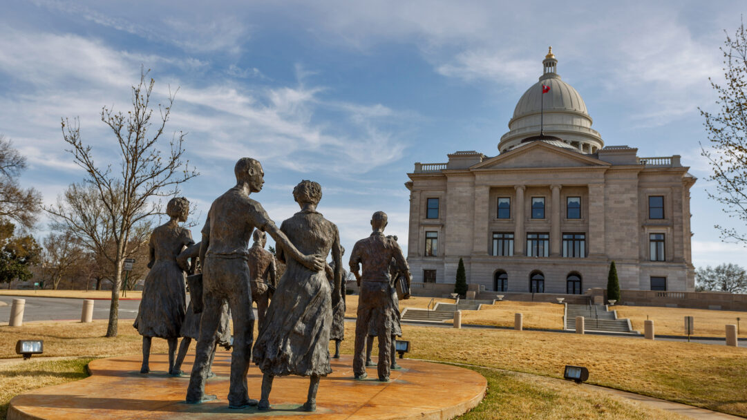 A poignant 4K wallpaper displaying the Testament The Little Rock Nine Monument in the foreground, with the grand Arkansas State Capitol Building rising majestically in the background, set in Little Rock under a bright sky. The bronze figures of the monument are sharply defined against the Capitol's stately facade, while dappled sunlight highlights the textures of the statues and the golden winter grass, evoking a sense of solemn history and enduring resolve.