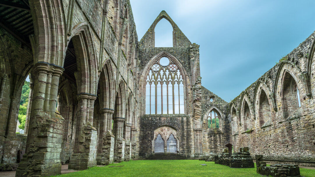 A majestic 4K wallpaper showcases the dramatic Gothic architecture of Tintern Abbey, revealing its ancient ruins set against a serene sky in Wales. Towering stone arches and a grand, open window frame dominate the view, with lush green grass filling the abbey's central space, emphasizing its timeless beauty and tranquil decay.