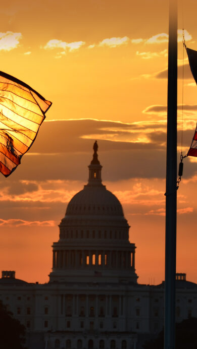 A breathtaking 4K wallpaper captures the US Capitol Building and two American flags at sunset in Washington DC. The deep orange and yellow sky dramatically silhouettes the historic landmark and illuminates the flags from behind, creating a patriotic and serene mood.