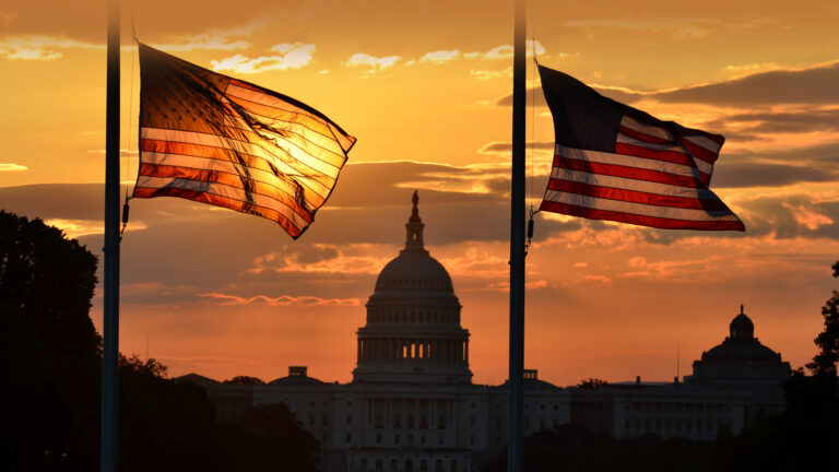 A breathtaking 4K wallpaper captures the US Capitol Building and two American flags at sunset in Washington DC. The deep orange and yellow sky dramatically silhouettes the historic landmark and illuminates the flags from behind, creating a patriotic and serene mood.