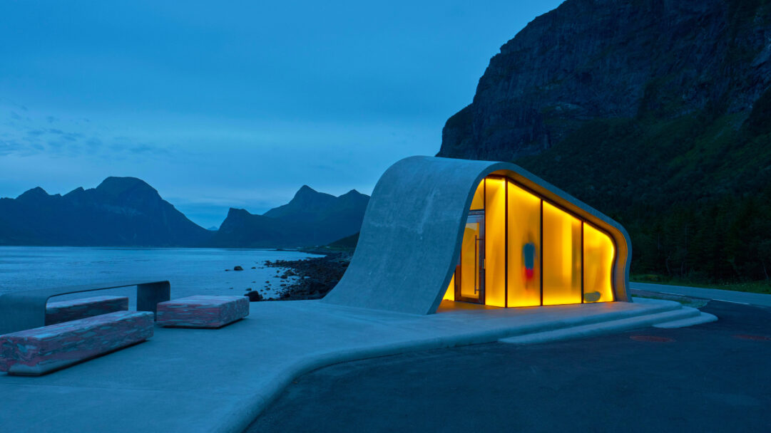 A captivating 4K wallpaper of the modern Ureddplassen Rest Area at night, nestled along Norway's Helgelandskysten Scenic Route. Its distinctive concrete structure glows with warm orange light against the serene blue twilight, reflecting softly on the calm water and silhouetted mountains.