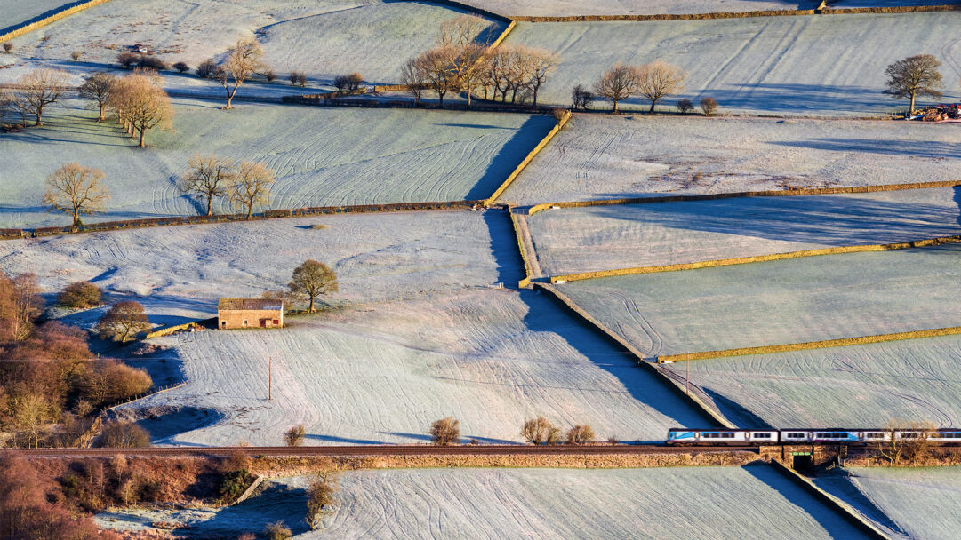 A serene 4K wallpaper showcasing a winter frost landscape in the Vale of Edale, Peak District England, where a train traverses the scenic terrain. The widespread frost blankets the patchwork fields in a delicate white, sharply defined by the long, cool blue shadows stretching across the land from bare trees and stone walls under the crisp winter sun.