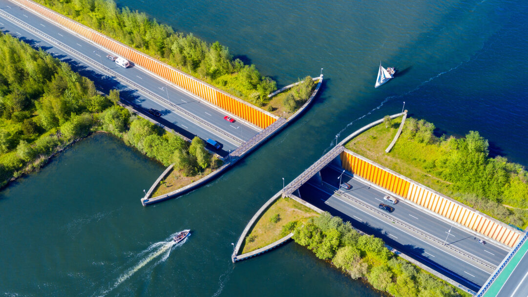 An ingenious 4K wallpaper presenting the Veluwemeer Aqueduct in the Netherlands, viewed from an aerial perspective, showcasing a busy road traveling beneath a body of water. Cars and caravans traverse the vivid gray road, while various boats, including a sailboat and a motorboat, glide over the deep blue aqueduct channel, creating a harmonious blend of land and water travel.