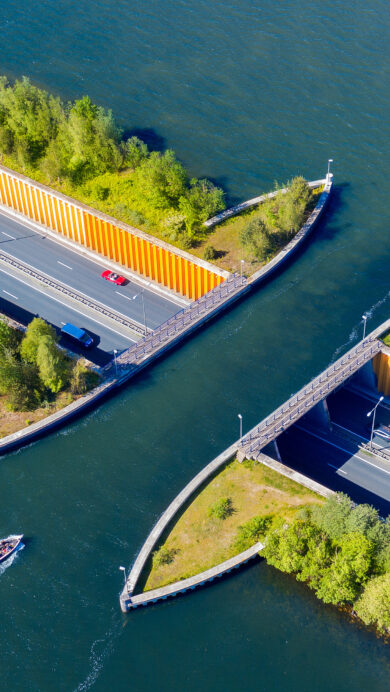 An ingenious 4K wallpaper presenting the Veluwemeer Aqueduct in the Netherlands, viewed from an aerial perspective, showcasing a busy road traveling beneath a body of water. Cars and caravans traverse the vivid gray road, while various boats, including a sailboat and a motorboat, glide over the deep blue aqueduct channel, creating a harmonious blend of land and water travel.