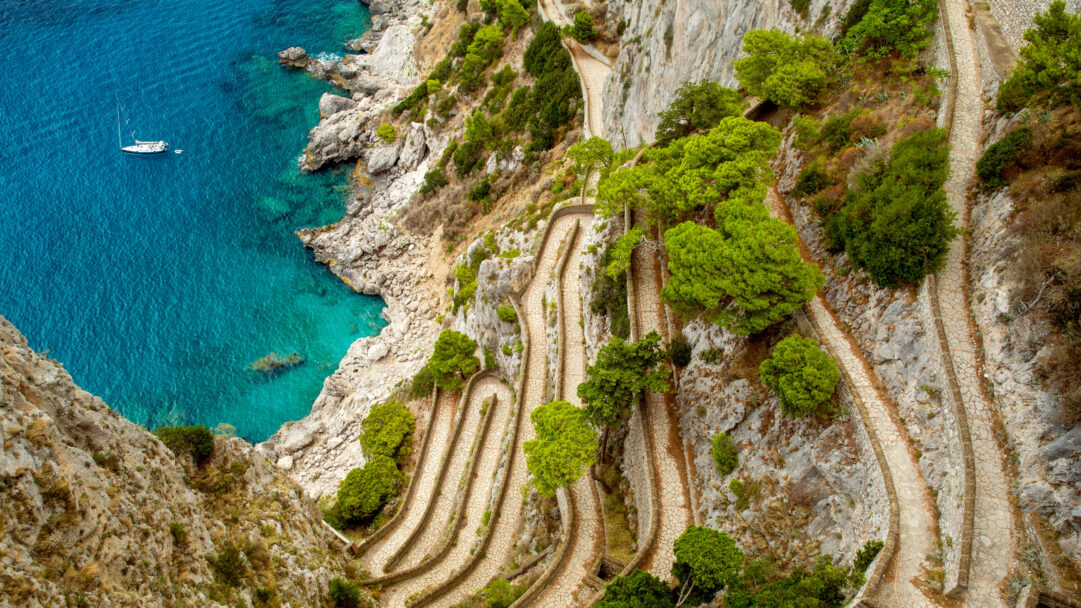 A breathtaking 4K wallpaper showcasing the iconic Via Krupp Footpath winding down the steep, rocky cliffs of Capri Island, Italy, overlooking the vibrant turquoise Mediterranean Sea. Its distinct serpentine curves are bordered by lush green foliage and rugged stone, contrasting beautifully with the sparkling clear water where a solitary sailboat glides, evoking a serene and adventurous coastal mood.