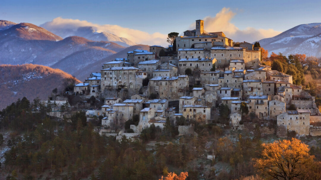 An enchanting 4K wallpaper displaying the historic Village of Labro, a winter hilltop settlement in Italy's Rieti Province, where stone buildings are lightly dusted with snow. Golden sunlight dramatically illuminates the village's ancient architecture and distant snowy mountains, creating a serene contrast with the cool blue sky and soft, bright clouds.