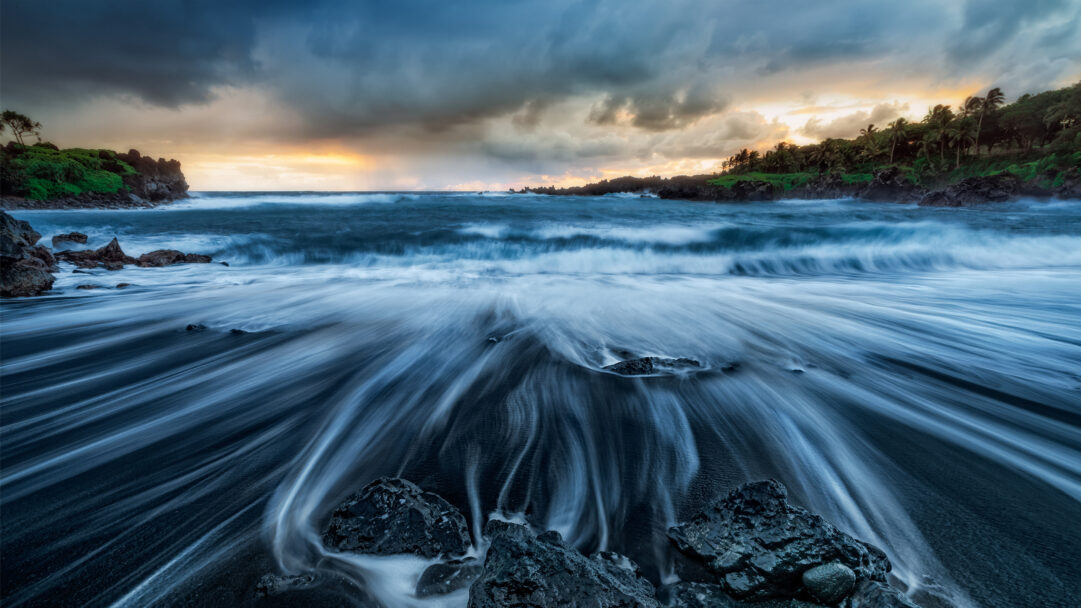A breathtaking 4K wallpaper capturing the Wai'ānapanapa Black Sand Beach in Maui, Hawaii, where powerful ocean waves meet the dark shore. Long exposure photography transforms the incoming tide into ethereal, milky streaks of blue and white flowing across the volcanic sand, creating a tranquil yet dynamic mood under a cloud-streaked sky with a golden sunset glow.