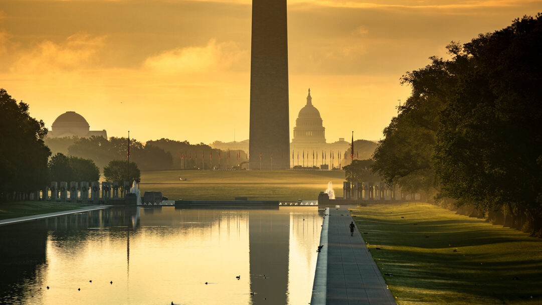 A radiant 4K wallpaper presenting the Washington Monument standing tall on the National Mall, with the Capitol Building visible in the hazy distance. The golden glow of sunrise beautifully illuminates the scene, casting a perfect reflection of the Monument and sky in the tranquil water below.