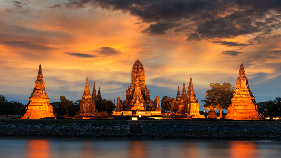 A breathtaking 4K wallpaper featuring Wat Chaiwatthanaram Temple in Ayutthaya, Thailand, dramatically illuminated at sunset beside a calm river. The ancient structures glow with the warm oranges and golds of the sunset, casting a luminous reflection on the tranquil water below, creating a serene and majestic ambiance.
