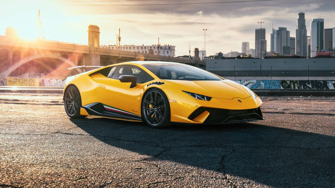 A breathtaking 4K wallpaper showcasing a radiant yellow Lamborghini Huracan parked on an asphalt street at sunset, with a prominent city skyline and an arched bridge in the background. The intense golden hour sunlight bathes the car and foreground in a warm glow, creating dramatic shadows and highlighting the vehicle's vibrant color against the urban backdrop.