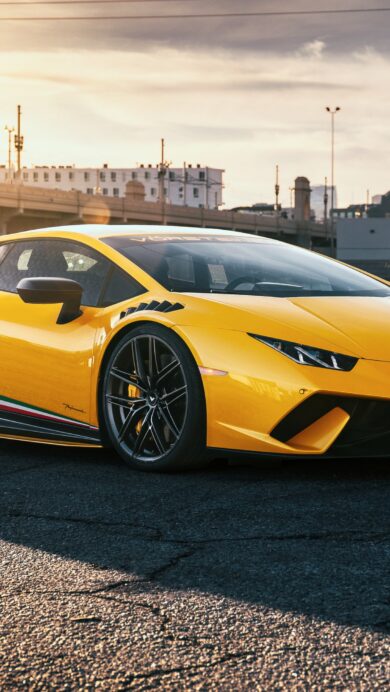 A breathtaking 4K wallpaper showcasing a radiant yellow Lamborghini Huracan parked on an asphalt street at sunset, with a prominent city skyline and an arched bridge in the background. The intense golden hour sunlight bathes the car and foreground in a warm glow, creating dramatic shadows and highlighting the vehicle's vibrant color against the urban backdrop.