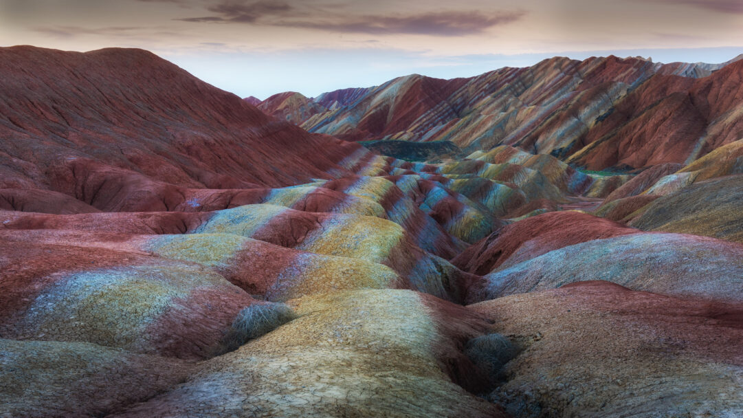 An awe-inspiring 4K wallpaper presents the iconic Rainbow Mountains landscape of Zhangye Danxia National Geopark. Striking geological formations showcase incredible stratified bands of red, orange, yellow, and blue, creating a vibrant, otherworldly spectacle under the soft, muted sky.
