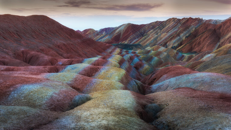 An awe-inspiring 4K wallpaper presents the iconic Rainbow Mountains landscape of Zhangye Danxia National Geopark. Striking geological formations showcase incredible stratified bands of red, orange, yellow, and blue, creating a vibrant, otherworldly spectacle under the soft, muted sky.
