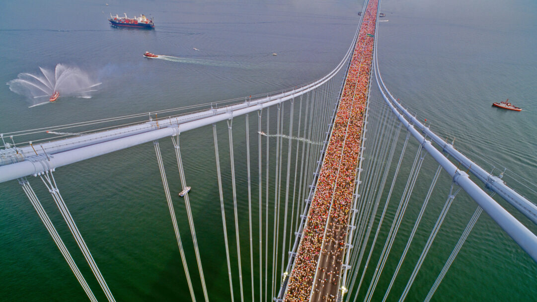An exhilarating 4K wallpaper captures thousands of New York City Marathon runners in 1990, streaming across the iconic Verrazzano-Narrows Bridge from an aerial perspective. The vibrant human river of athletes vividly contrasts with the monumental grey suspension cables and the deep blue-green waters below, where celebratory fireboats spray impressive arcs of white.