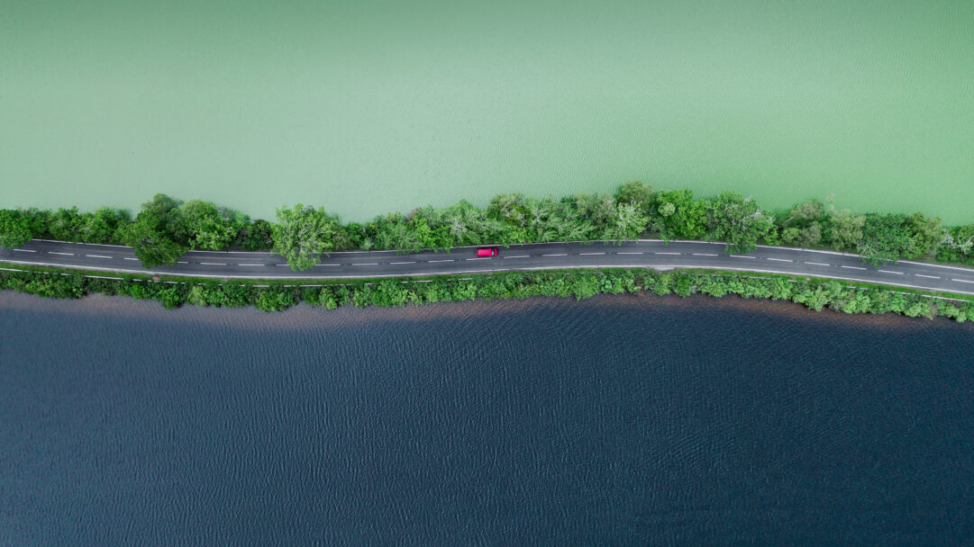 A breathtaking 4K wallpaper of an aerial view showing a narrow road, lined with lush green trees, neatly separating a vibrant light green lake from a deep blue one in the Scottish Highlands. The stark contrast between the vivid green and dark blue waters, sharply divided by the slender, tree-lined road with a solitary red car, creates a dramatic and balanced natural landscape.