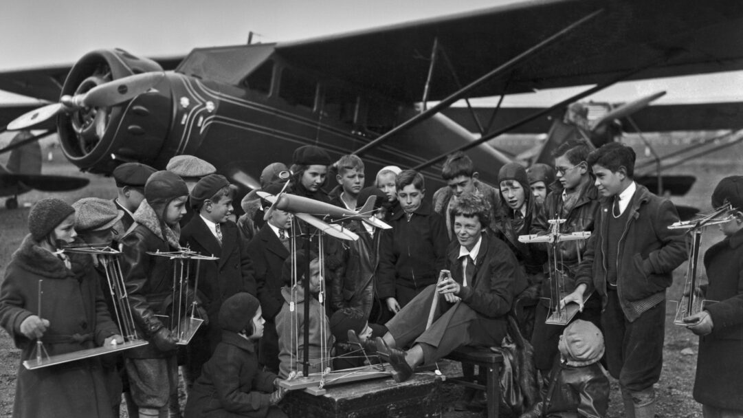 A captivating 4K wallpaper showcasing Amelia Earhart seated on an airfield in Newark, New Jersey, actively teaching a group of young aviation students in front of a substantial propeller plane. The black and white image highlights the eager faces of the students, many holding small model aircraft or instructional tools, engrossed in the lesson from the pioneering pilot.