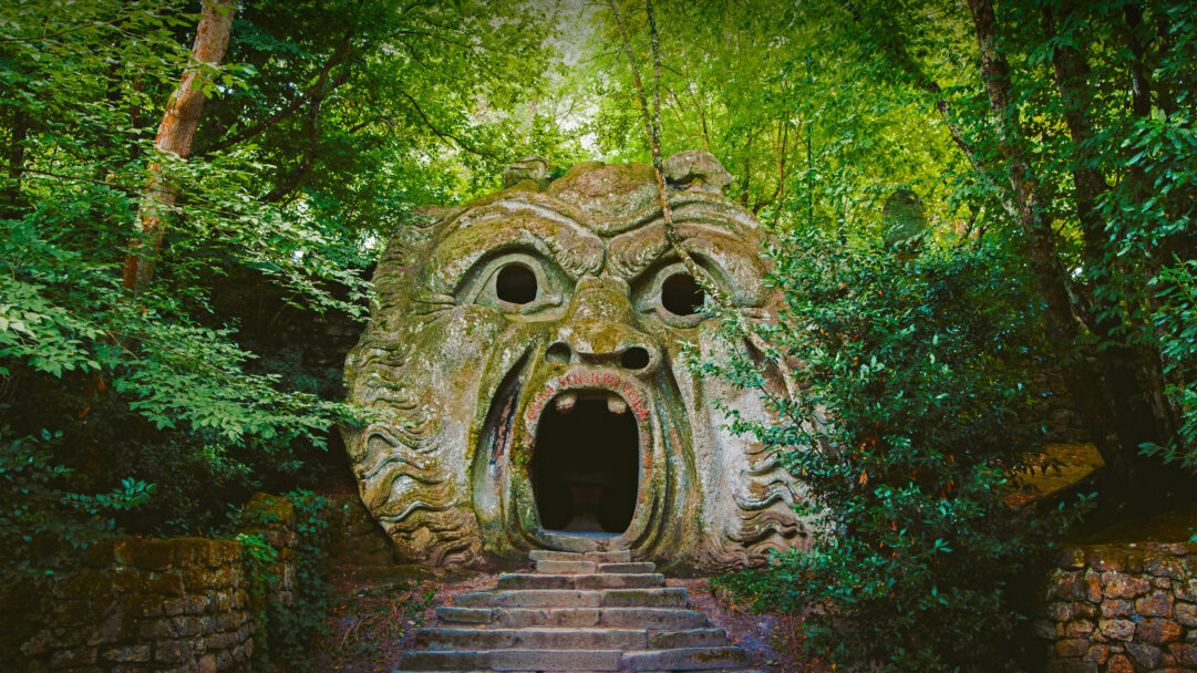 A haunting 4K wallpaper featuring the ancient Orcus stone face sculpture, deeply carved and nestled within the verdant, wild Gardens of Bomarzo, Italy. The sculpture's immense, weathered stone face, with its wide, cavernous mouth forming a passageway, emanates an ancient, powerful emotion amidst the surrounding dense green foliage.