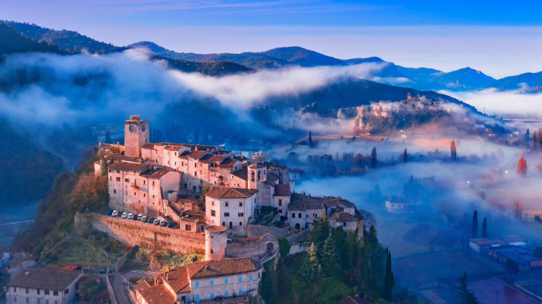 A breathtaking 4K wallpaper presenting Arrone Village in Umbria, Italy, nestled majestically within a sprawling mountain landscape. Golden morning light illuminates the ancient stone buildings of the village, dramatically contrasting with the swirling, cool blue fog that blankets the valleys and distant mountains.