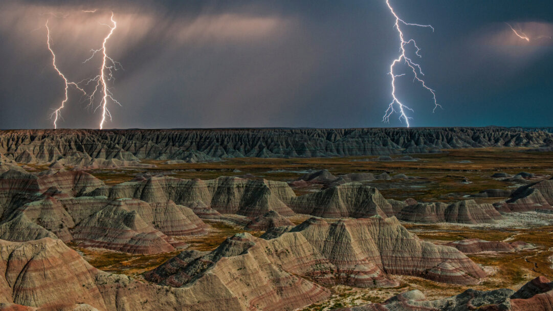 An awe-inspiring 4K wallpaper captures the dramatic landscape of Badlands National Park in South Dakota, showcasing its distinctive rock formations under a tumultuous sky. Two spectacular lightning bolts violently crack through the dark clouds, vividly highlighting the rugged, layered striations of the hills and intensifying the raw power of the storm.