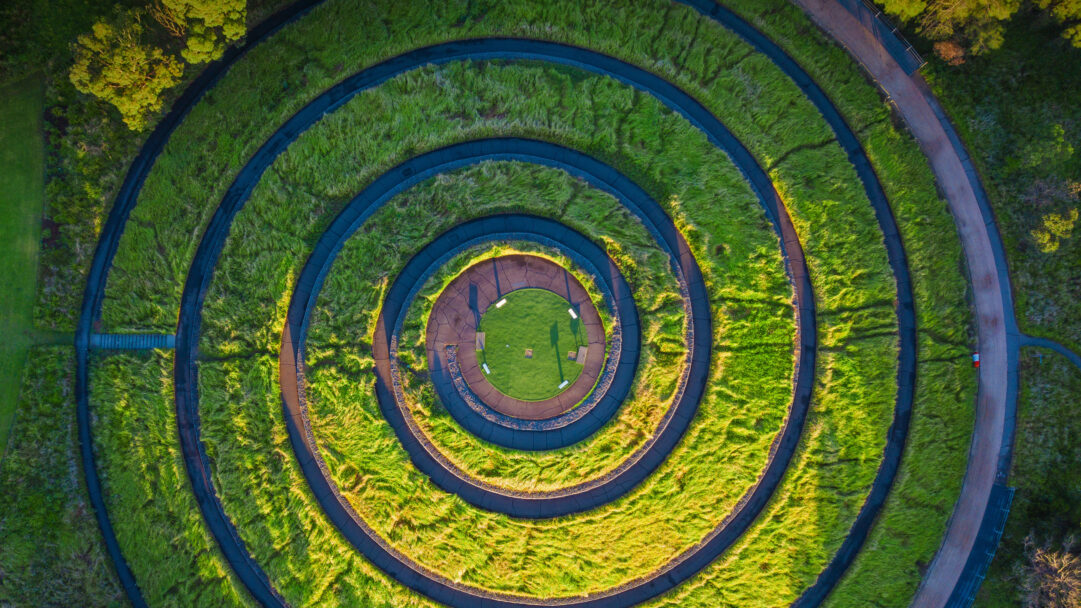 A geometrically striking 4K wallpaper presenting an aerial view of the Bay Marker Lookout in Sydney Olympic Park, Australia, composed of concentric circular green pathways. Vividly illuminated by sunlight, the contrasting dark paved paths spiral through undulating bands of bright green grass, creating a mesmerizing, almost hypnotic pattern.