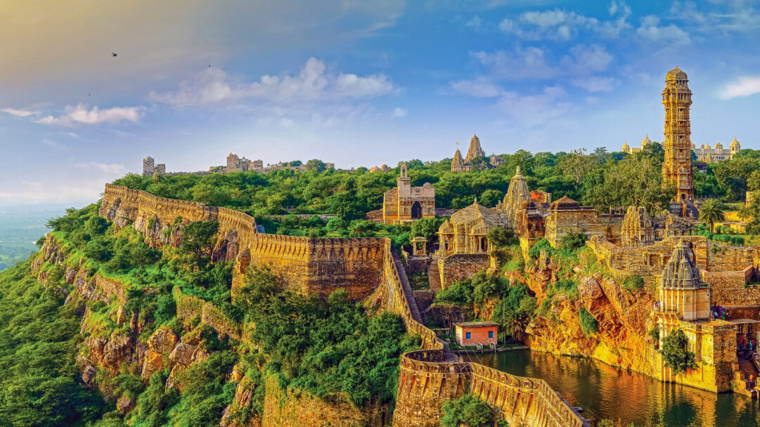 A captivating 4K wallpaper of the sprawling Chittorgarh Fort in India, an historic hilltop complex nestled amidst lush green hills under a bright sky. Golden sunlight illuminates the ancient stone walls and ornate temples, casting rich hues that highlight the complex's majestic scale and timeless beauty.