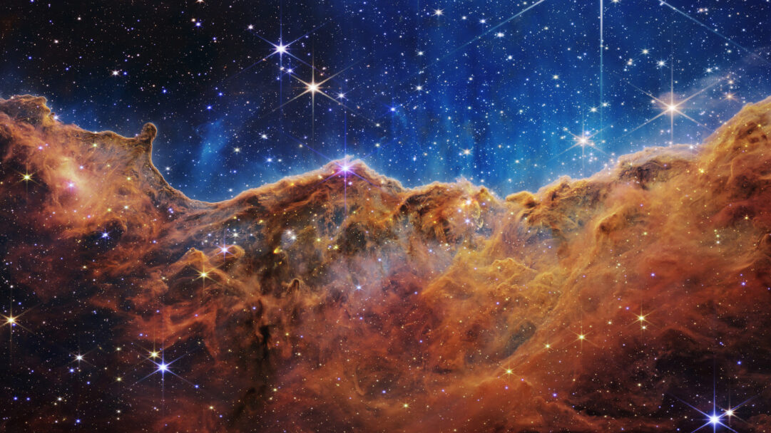 A breathtaking 4K wallpaper revealing the majestic Cosmic Cliffs of the Carina Nebula, a towering landscape of interstellar gas and dust within a star-studded expanse of deep space. Vibrant pillars of glowing orange and red nebula rise dramatically against a profound blue-black sky, punctuated by countless brilliant, distant stars that evoke a sense of immense cosmic creation.