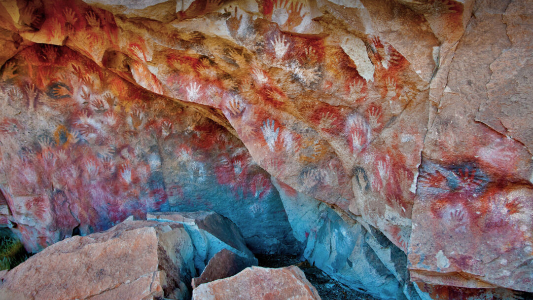 A breathtaking 4K wallpaper capturing a vast array of ancient handprints adorning the textured rock walls within the historic Cueva de las Manos cave in Santa Cruz, Argentina. The myriad handprints in striking reds, ochres, whites, and blacks dynamically cover the rough cave surfaces, creating a vivid historical tapestry bathed in soft, inviting light.