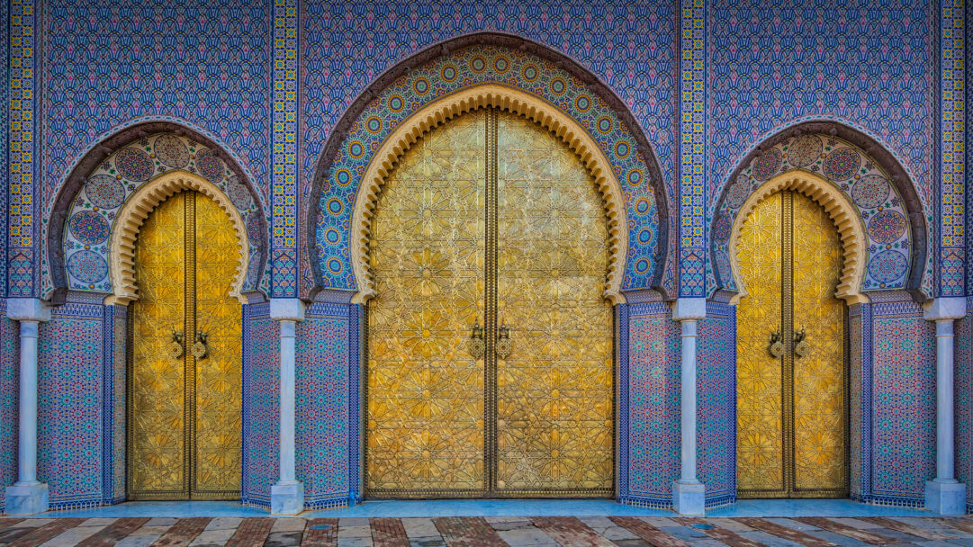 A breathtaking 4K wallpaper showcasing the iconic golden gates and intricate mosaic arches of the Royal Palace in Fez, Morocco. The dazzling gold of the ornate gates contrasts brilliantly with the vibrant blue and multicolored geometric patterns of the surrounding mosaic tiles, creating an atmosphere of regal splendor.