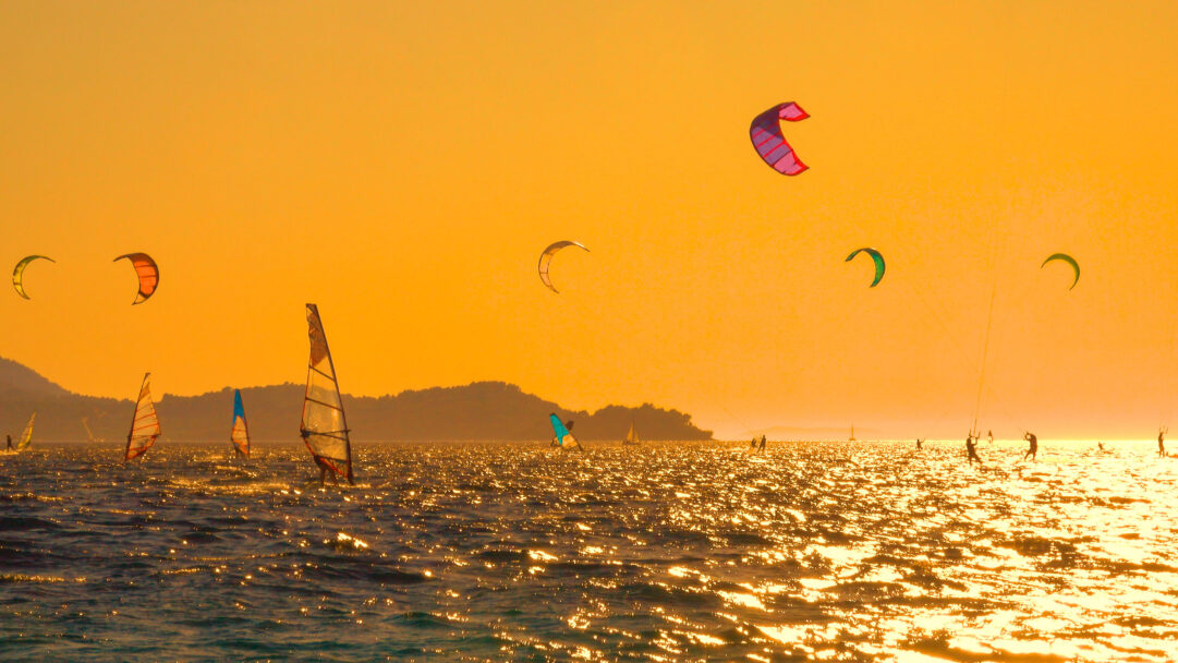 A breathtaking 4K wallpaper captures multiple kiteboarders and windsurfers actively enjoying the waters off Pelješac Peninsula, Croatia. The entire scene is bathed in the warm, intense glow of a golden sunset, making the water shimmer with countless bright reflections and silhouetting the dynamic figures against the radiant sky.