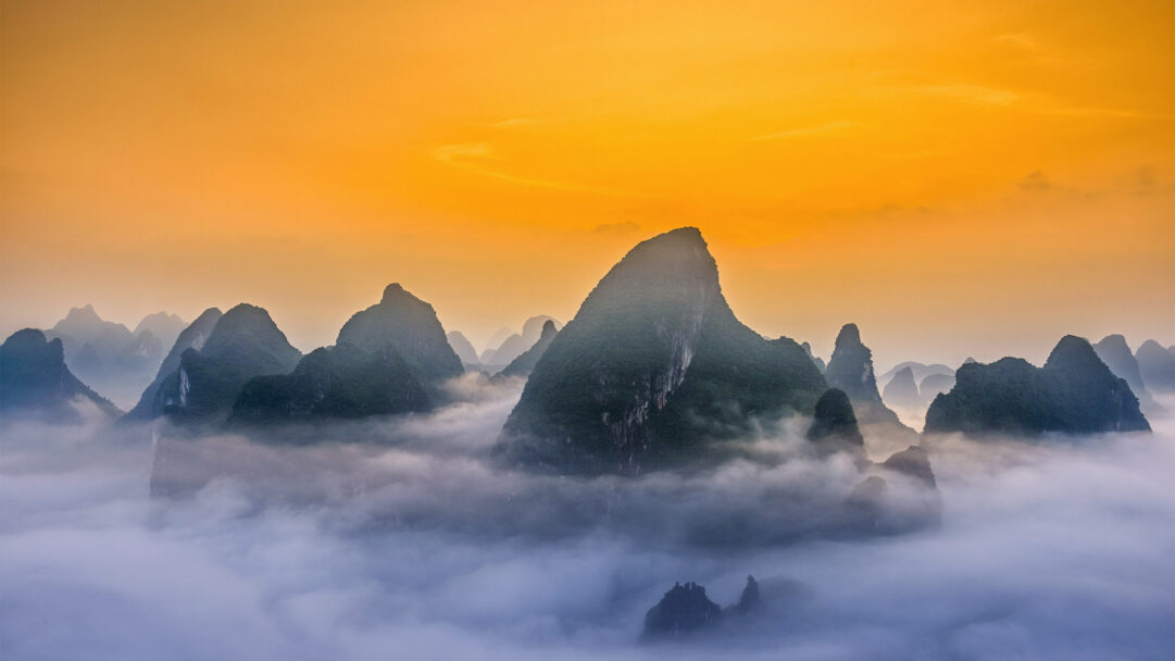An ethereal 4K wallpaper captures the iconic Guilin Lijiang River Karst Mountains rising majestically through a thick, undulating fog. The vibrant golden-orange hues of the sunrise illuminate the sky, casting a dramatic glow over the dark, forested peaks that pierce through the serene, smoky expanse.