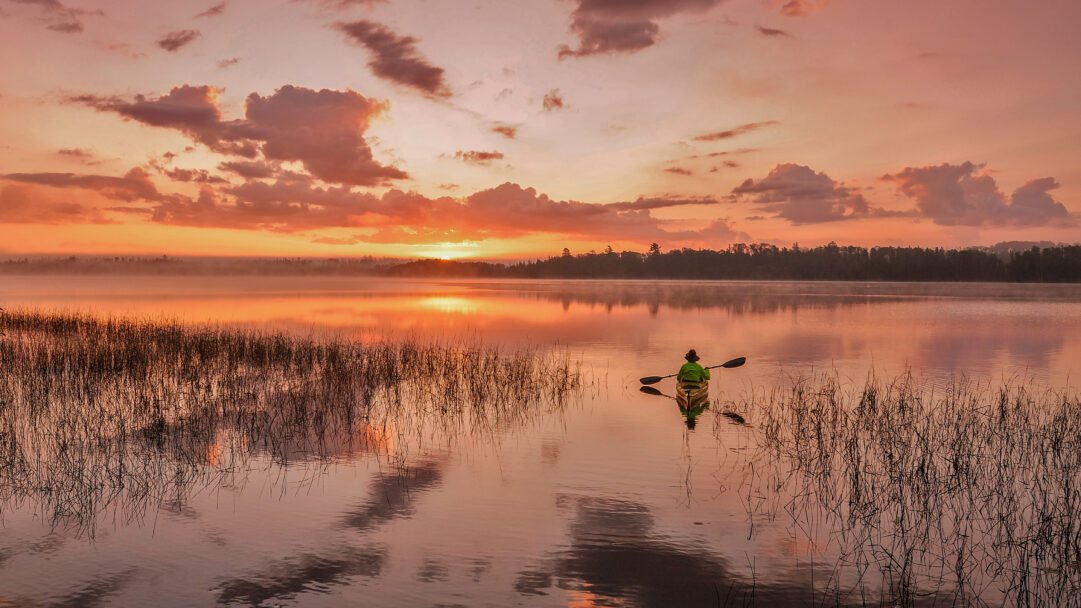 A tranquil 4K wallpaper showcasing a kayaker paddling across a foggy lake at sunset in the Boundary Waters Canoe Area Wilderness, Minnesota. The sky blazes with fiery oranges and soft pinks, casting a mirror-like reflection on the misty water, enveloping the scene in a serene glow.