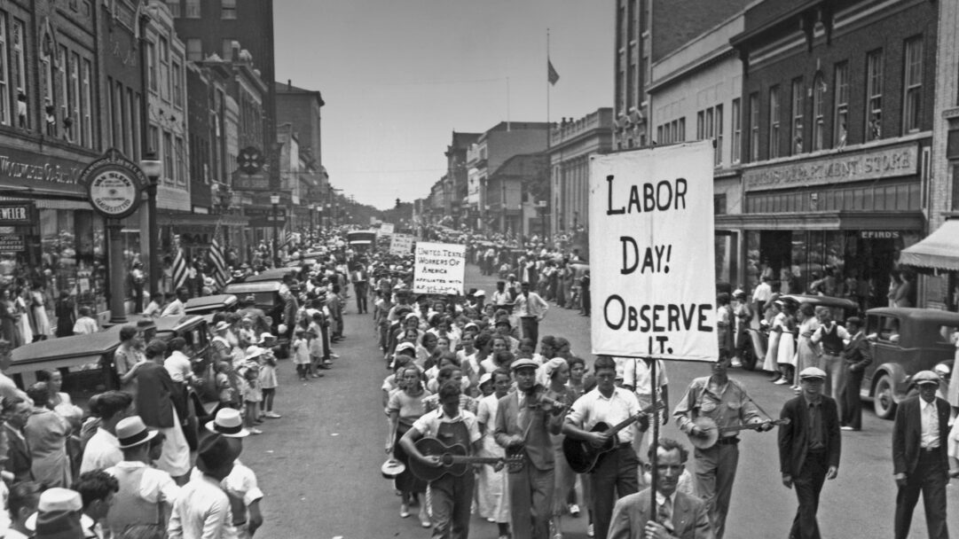 A compelling 4K wallpaper depicts a vintage black and white scene of the 1934 Labor Day Parade making its way down a bustling street in Gastonia, North Carolina, flanked by onlookers and historic brick buildings. Scores of men, women, and children march, some playing guitars and fiddles, others carrying banners proclaiming Labor Day! Observe It. and United Textile Workers of America, evoking a sense of community and determination during the Great Depression era.