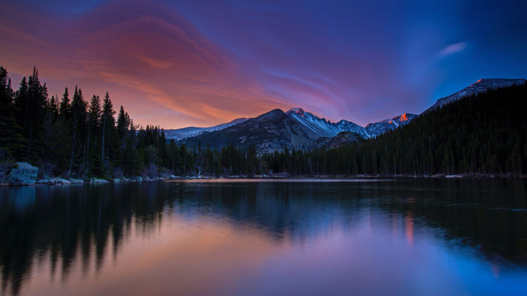 A sublime 4K wallpaper depicting Longs Peak, illuminated by alpenglow, rising majestically above a placid lake in Rocky Mountain National Park. The sky's dramatic purple and orange hues are flawlessly mirrored in the calm water, emphasizing the mountain's radiant peak and the dark evergreen forest lining the shore.
