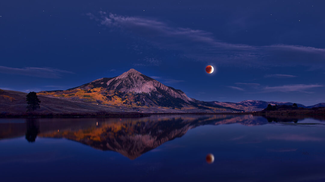 A mesmerizing 4K wallpaper captures a dramatic lunar eclipse glowing above Mount Crested Butte, Colorado, with its detailed reflection mirrored perfectly in a tranquil lake. The deep reddish-orange hue of the eclipsed moon and its reflection contrast beautifully with the serene blue twilight sky and the golden autumn foliage on the mountain slopes, creating a captivating and otherworldly mood.