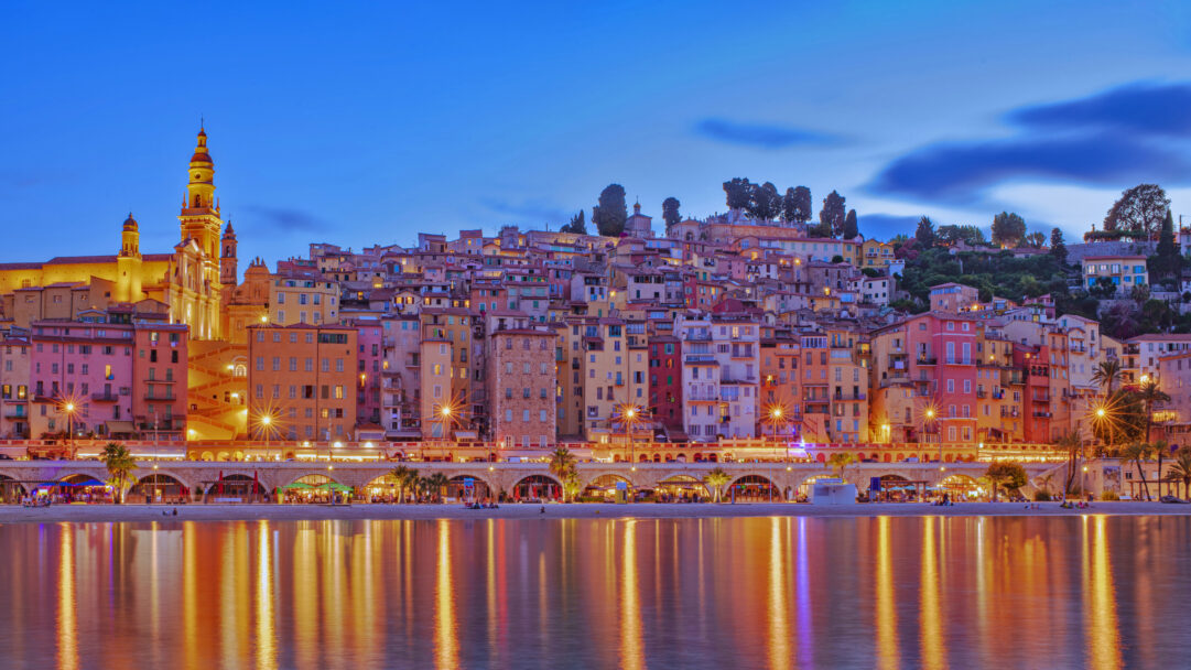 An enchanting 4K wallpaper of the Menton, France evening cityscape, where a hillside of colorful buildings descends to a tranquil waterfront. The vibrant lights from the buildings and the illuminated church tower cast brilliant, elongated reflections across the calm water, capturing the city's lively yet serene evening mood.