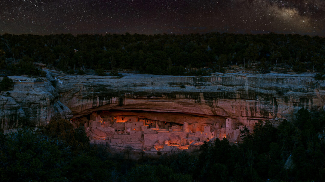 An ethereal 4K wallpaper showcasing the ancient Mesa Verde National Park Cliff Dwellings nestled within a vast rock face at night. Warm, golden light glows from within the dwellings, casting an inviting hue against the cool tones of the cliff and the brilliant, star-strewn Milky Way overhead.