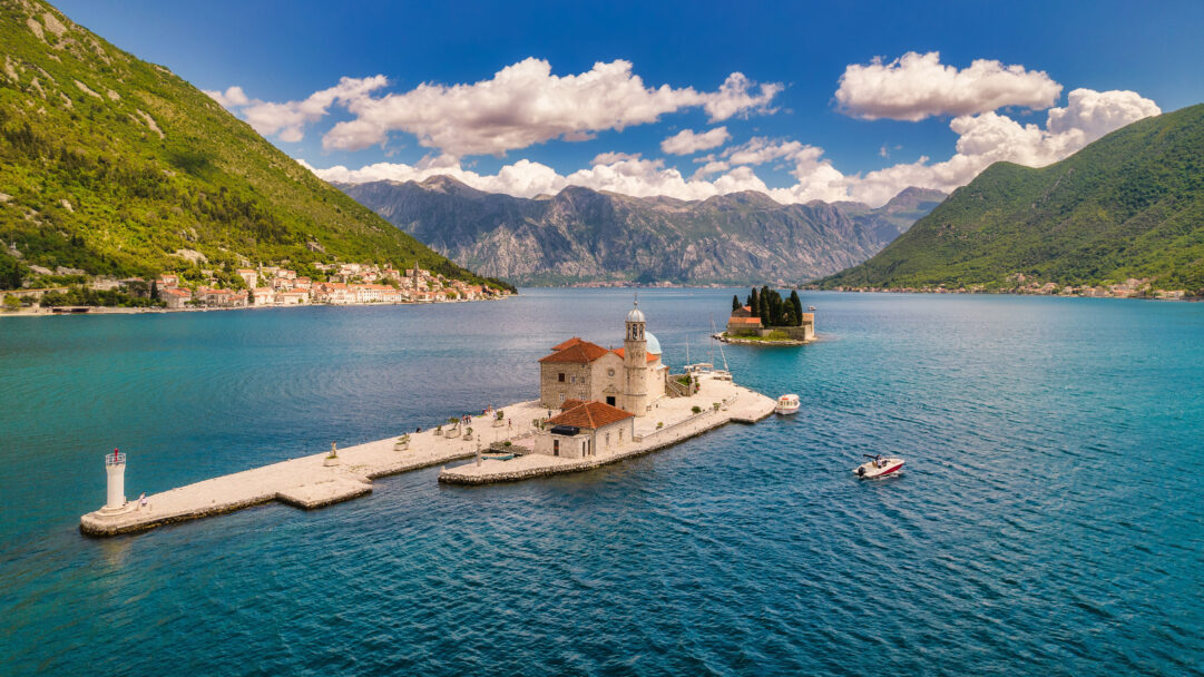 A breathtaking 4K wallpaper capturing the historic Our Lady of the Rocks and Saint George Island nestled within Montenegro's beautiful Bay of Kotor, with the charming town of Perast lining the left shore. The serene turquoise waters of the bay reflect the towering green mountains and a dramatic blue sky, creating a vibrant scene of natural beauty and cultural heritage.