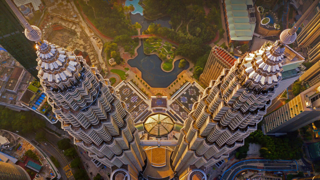 A magnificent 4K wallpaper presents the iconic Petronas Twin Towers in Kuala Lumpur, Malaysia, viewed from a striking overhead perspective. Warm sunlight dramatically illuminates the towers' intricate silver facades, casting long shadows over the sprawling urban landscape and lush green park beneath.
