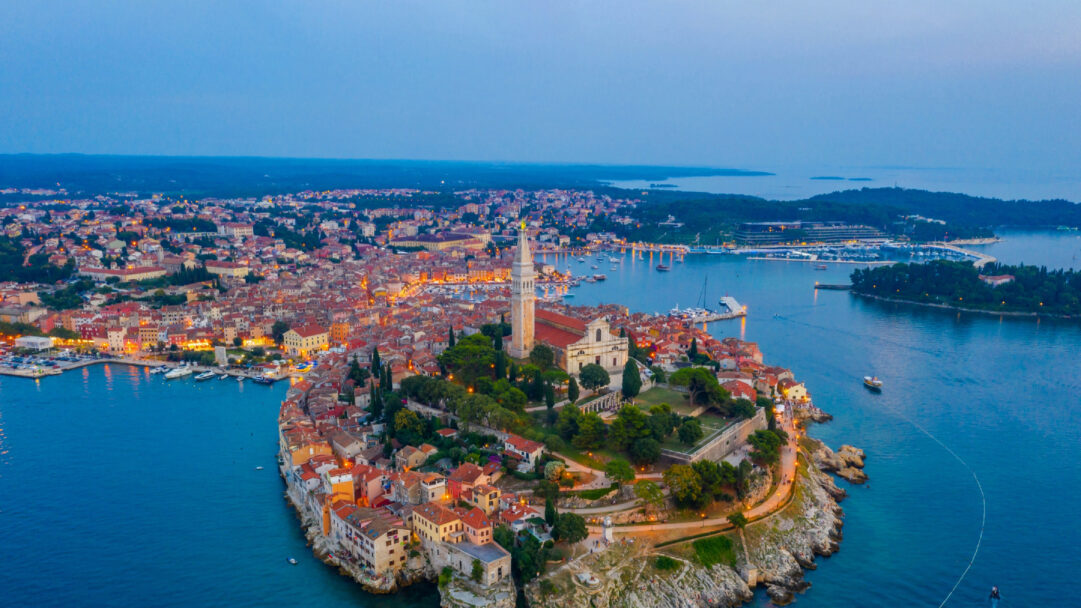 An enchanting 4K wallpaper capturing an aerial view of Rovinj Old Town in Croatia, a historic peninsula settlement dominated by a towering church and surrounded by the serene Adriatic Sea. At dusk, warm golden lights from the town's buildings and harbor begin to twinkle, casting soft reflections on the deep blue water, creating a tranquil and picturesque twilight scene.