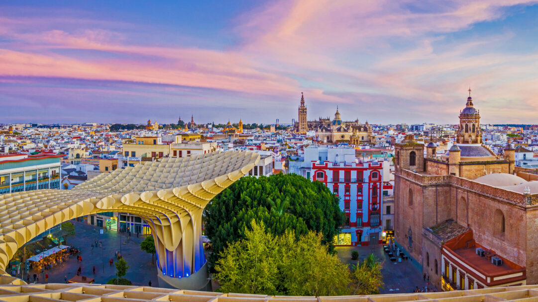 An enchanting 4K wallpaper presents an expansive Seville city view at sunset, centered around the striking Metropol Parasol. The sky above glows with vibrant streaks of pink, purple, and blue, casting a serene yet lively atmosphere over the historic cityscape and its modern architectural marvel.