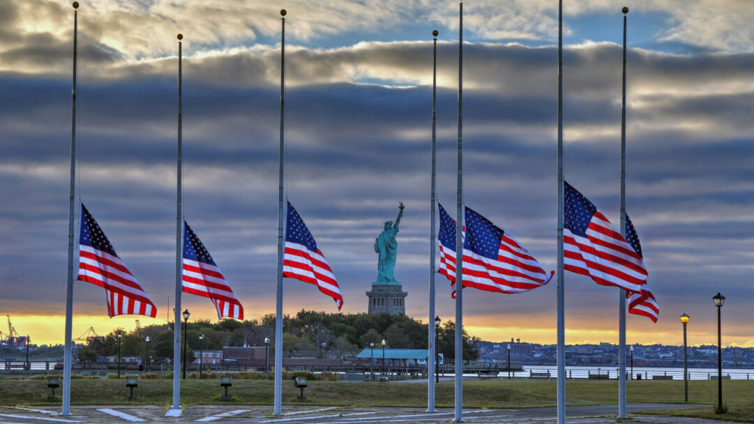 A solemn 4K wallpaper captures the Statue of Liberty overlooking New York City, flanked by multiple US flags flying at half-staff. The dramatic sky, with its blend of dark clouds and golden horizon, underscores the poignant mood of remembrance for the 9/11 Anniversary in 2014.