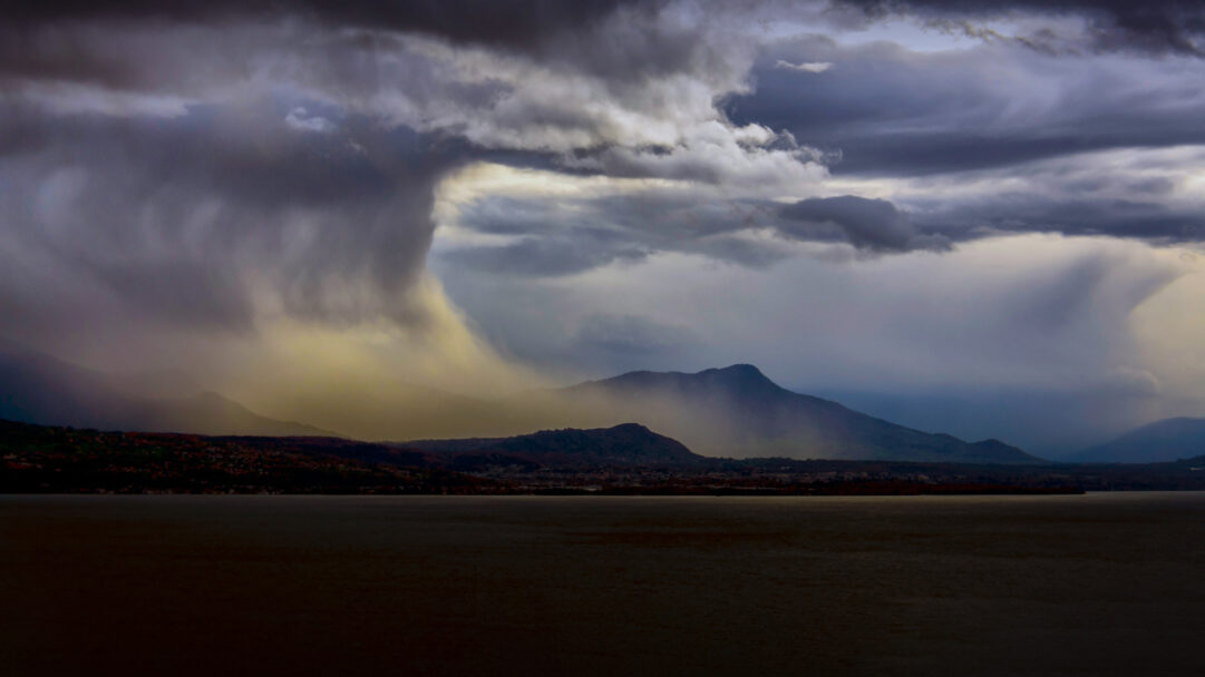 A powerful 4K wallpaper capturing a dramatic stormy sky descending over the vast expanse of Lake Geneva, with the silhouette of Lausanne and distant Swiss mountains visible along the shore. A colossal, dark rain shaft dominates the left, illuminated by an ethereal golden light, creating a moody and imposing atmosphere over the deep, reflective water.