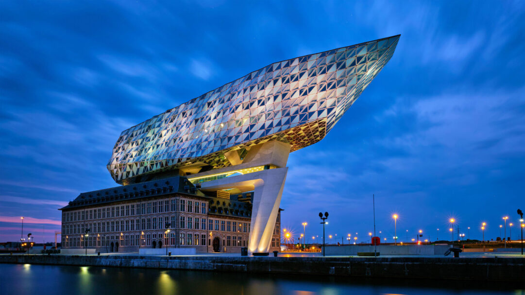 A dramatic 4K wallpaper of The Port House by Zaha Hadid Architects in Antwerp, Belgium, standing brilliantly illuminated against a deep blue night sky above tranquil port waters. Its shimmering, diamond-patterned glass extension, resembling a massive ship, casts a vibrant glow that contrasts sharply with the classic brick base and the city lights beyond.