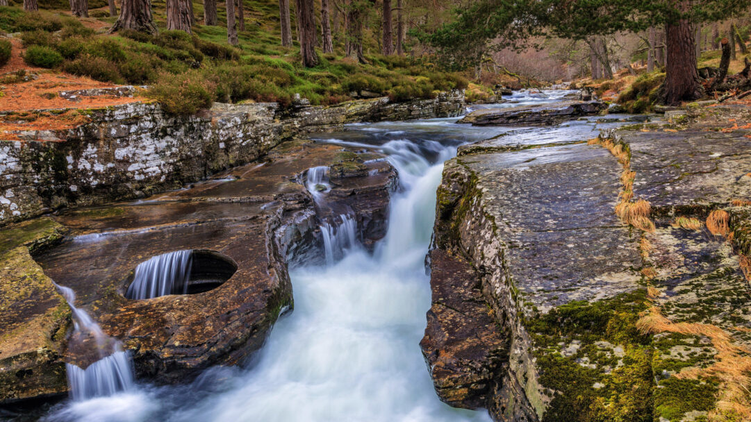 An immersive 4K wallpaper captures The Punch Bowl Waterfall on the River Quoich in the Cairngorms, Scotland, nestled within a lush forest setting. Its powerful white cascade, softened by a long exposure, dramatically plunges into dark, carved rock pools, creating a dynamic contrast with the rich, moss-covered banks and ancient pine trees.