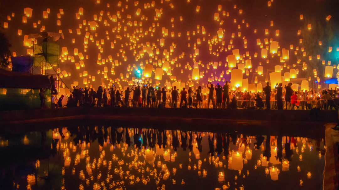 An enchanting 4K wallpaper captures the Yi Peng Festival in Chiang Mai, Thailand, at night, with thousands of luminous sky lanterns rising into the dark sky above a crowd gathered by a reflective body of water. The warm golden glow of the lanterns creates a dazzling spectacle, mirrored beautifully on the water's surface, evoking a sense of wonder and joyous celebration.