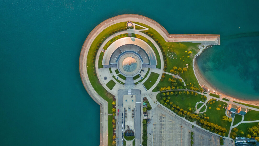 A captivating 4K wallpaper presents an aerial view of the Adler Planetarium in Chicago, Illinois, majestically situated on a peninsula extending into the vast, teal waters of Lake Michigan. The planetarium's distinct circular and semicircular architecture, surrounded by vibrant green lawns and winding pathways, glows with warm, golden sunlight, creating a striking contrast with the deep blue-green of the lake.
