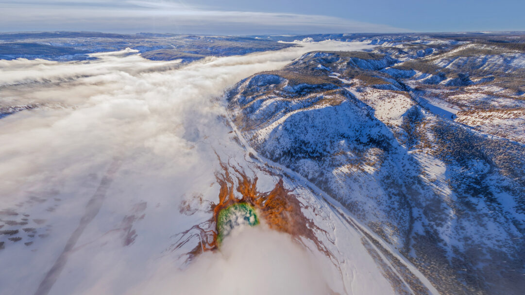 A dramatic 4K wallpaper revealing an aerial winter view of Grand Prismatic Spring in Yellowstone National Park, partly shrouded by a dense blanket of fog. The vibrant geothermal colors of the spring—fiery oranges and brilliant greens—pierce through the ethereal white mist, creating a striking contrast with the vast, snow-covered landscape.