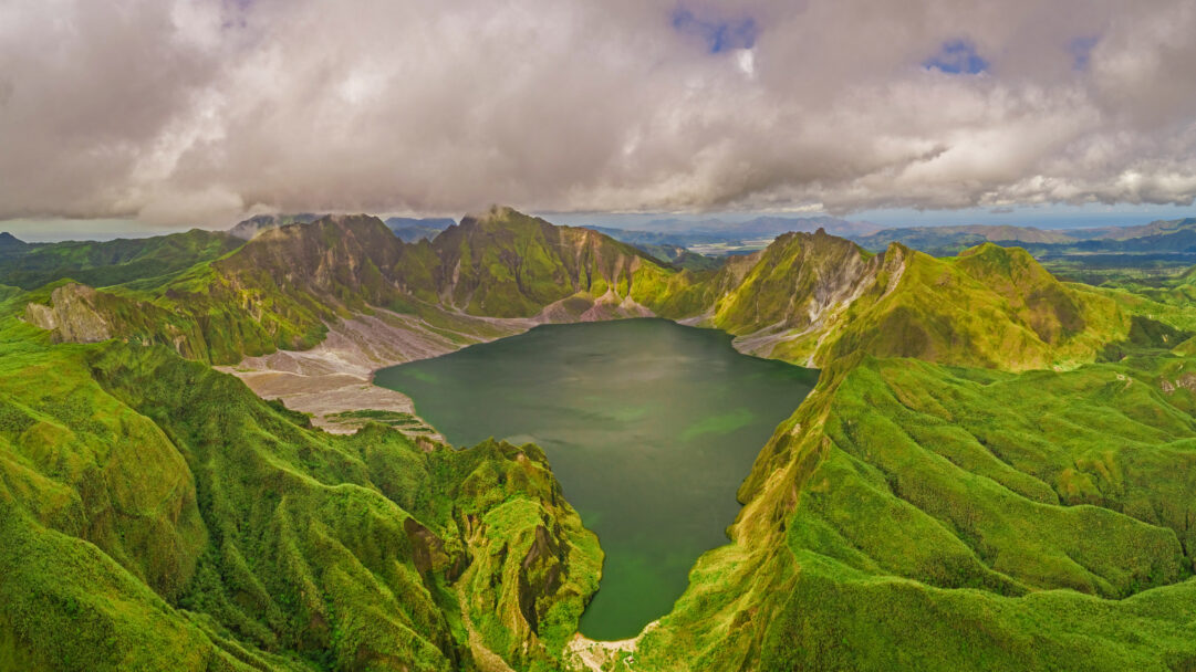 A breathtaking 4K wallpaper capturing an aerial view of the majestic Volcanic Lake Pinatubo, nestled amidst vast, lush green mountains under a dramatic, cloudy sky. The deep emerald green of the crater lake beautifully contrasts with the vibrant, intensely textured green slopes of the surrounding caldera, evoking a sense of raw natural power and serene grandeur.